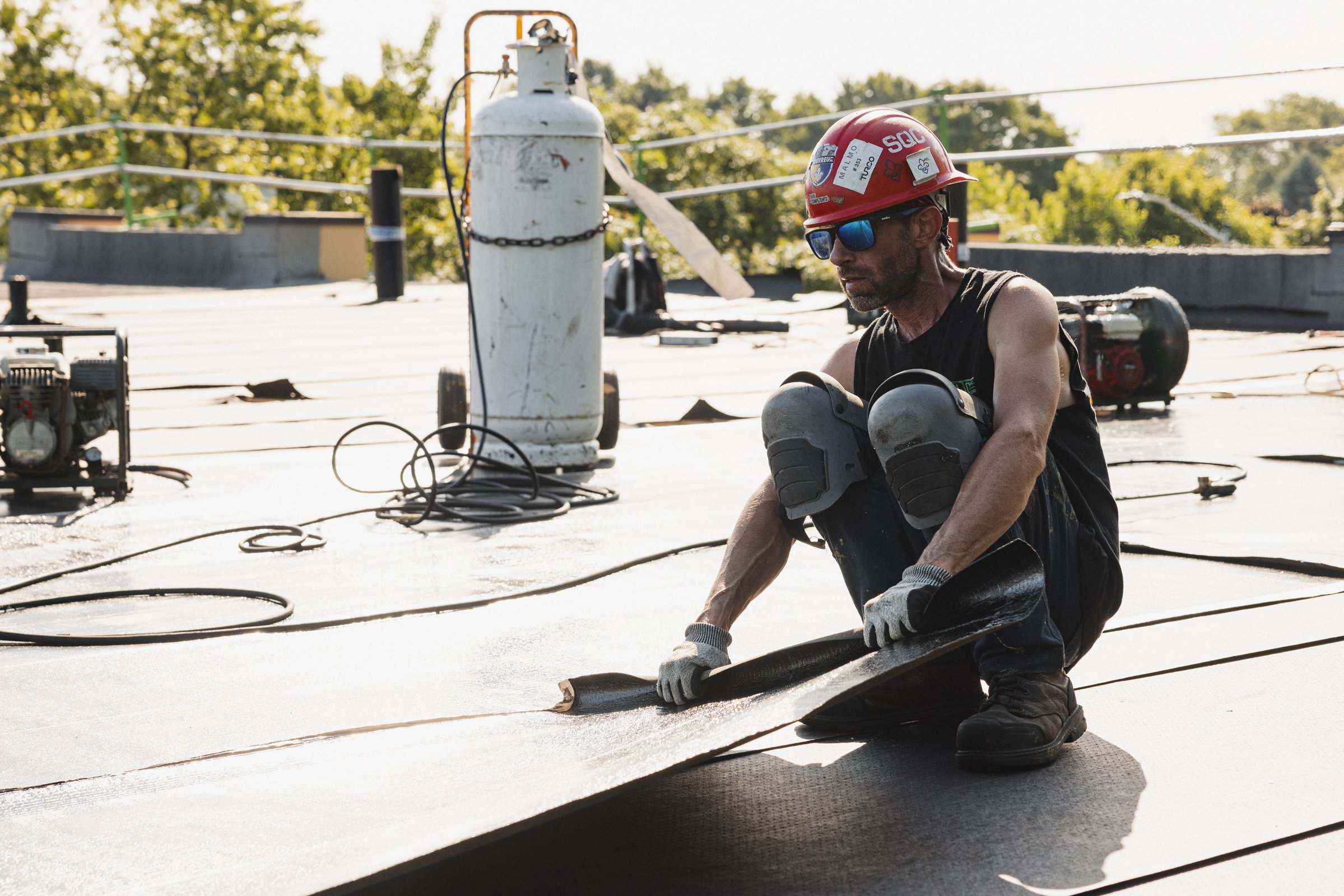 Un homme en tenue de travail, portant un casque rouge avec des autocollants et des lunettes de soleil, est à genoux sur une toiture en train de retirer un morceau de matériau de toiture, avec des outils et des équipements de toiture autour de lui.