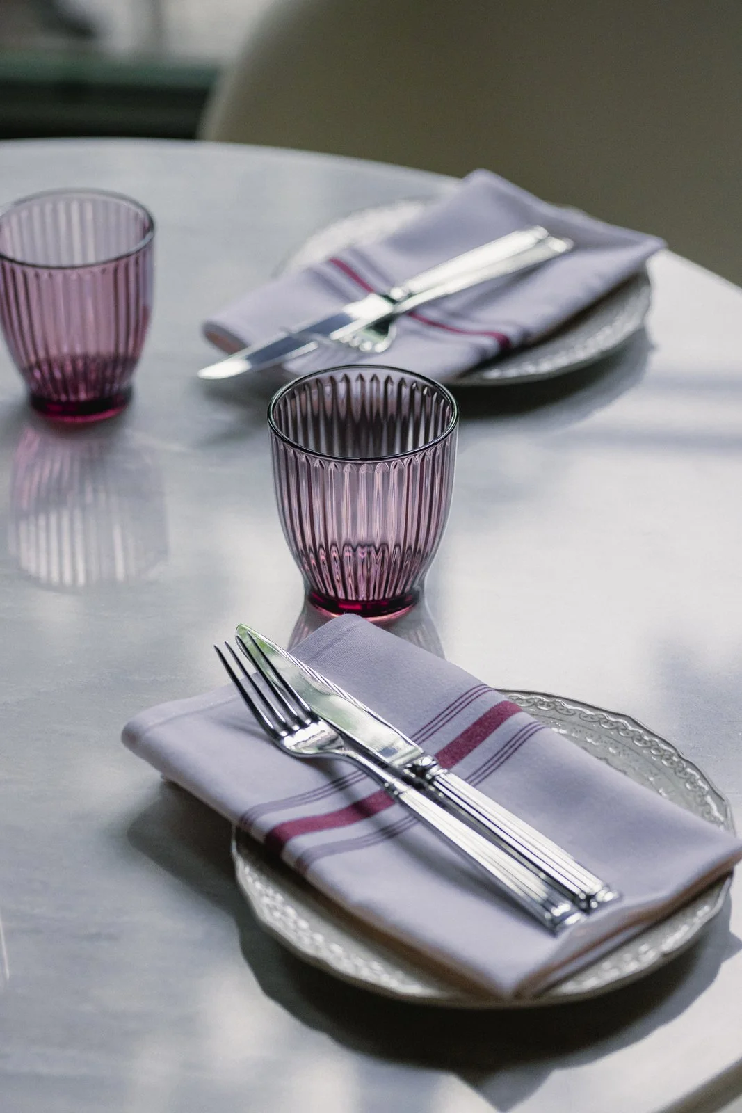 Une table dressée avec deux assiettes, des fourchettes, couteaux, verres en verre violet, et des serviettes blanches avec des rayures rouges.