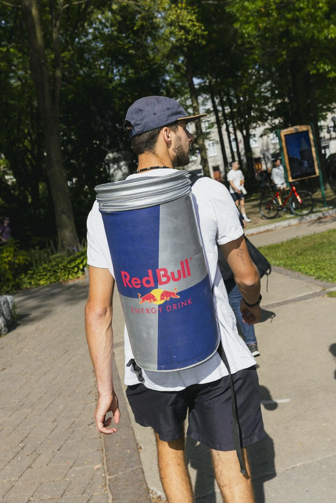 Jeune homme portant un sac à dos en forme de canette Red Bull dans un parc ensoleillé, avec des arbres et des personnes qui marchent en arrière-plan.