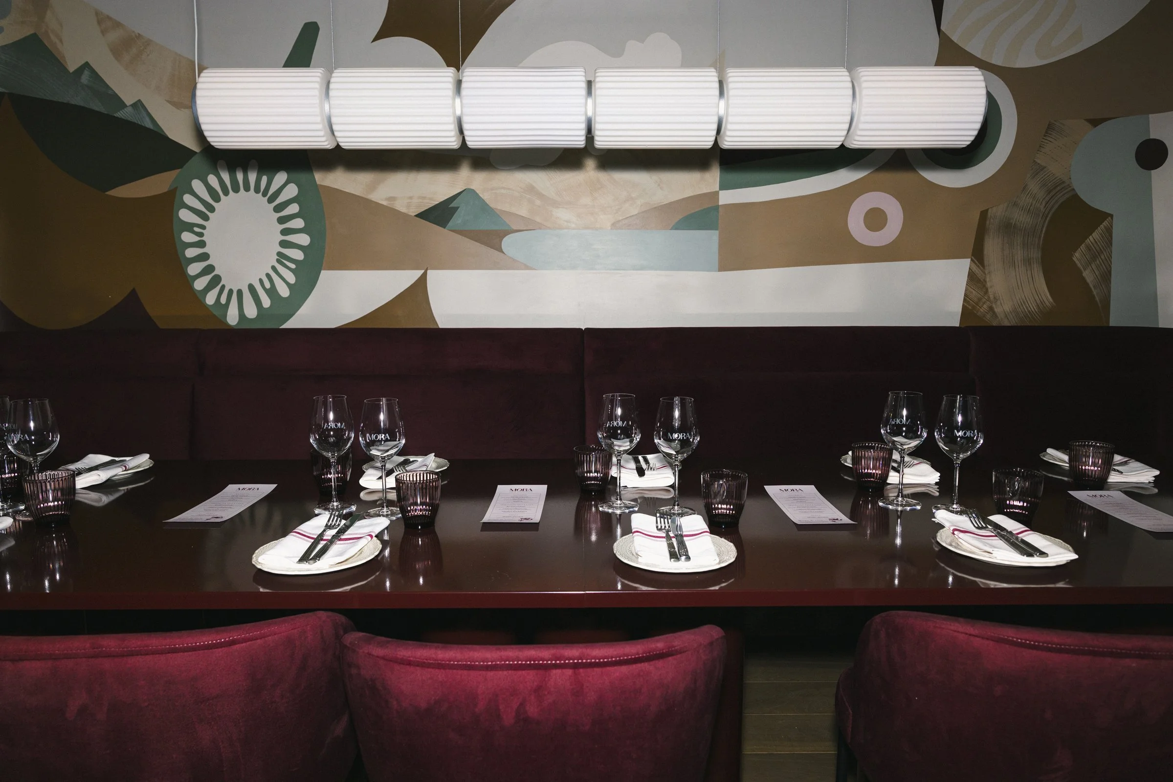 Table dressée dans un restaurant avec verres à vin, assiettes, couverts et menus, décorée avec un mur peint artistique en arrière-plan.