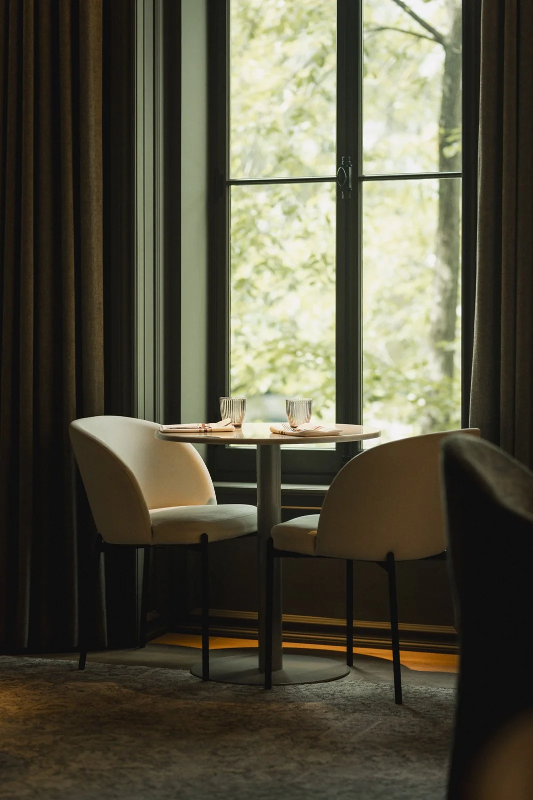 Une table ronde avec deux verres et des couverts, située devant une grande fenêtre laissant entrer de la lumière naturelle, dans un intérieur élégant et cosy.