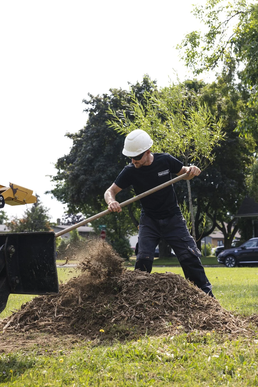 Un homme portant un casque de sécurité, des lunettes de soleil et un t-shirt noir utilise une pelle pour déplacer de la terre dans un jardin ou un parc, avec des arbres en arrière-plan et un ciel ensoleillé.