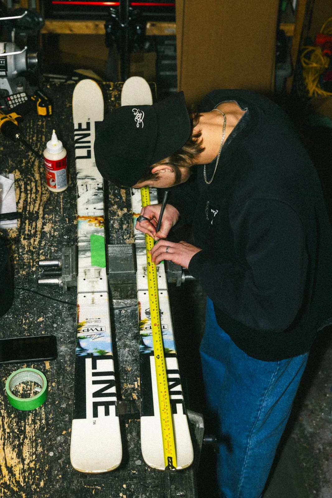 Une personne en train de mesurer des skis avec un ruban à mesurer sur un établi en bois. Plusieurs outils et produits sont visibles sur la table, dont de la colle et des tournevis.