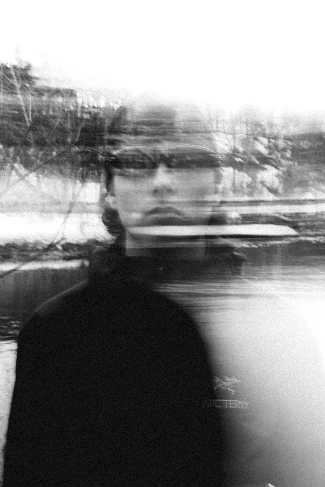 Une personne portant une veste noire avec un logo 'Arc'teryx' sur la poitrine, floue et doublement exposée, avec un paysage naturel en arrière-plan.