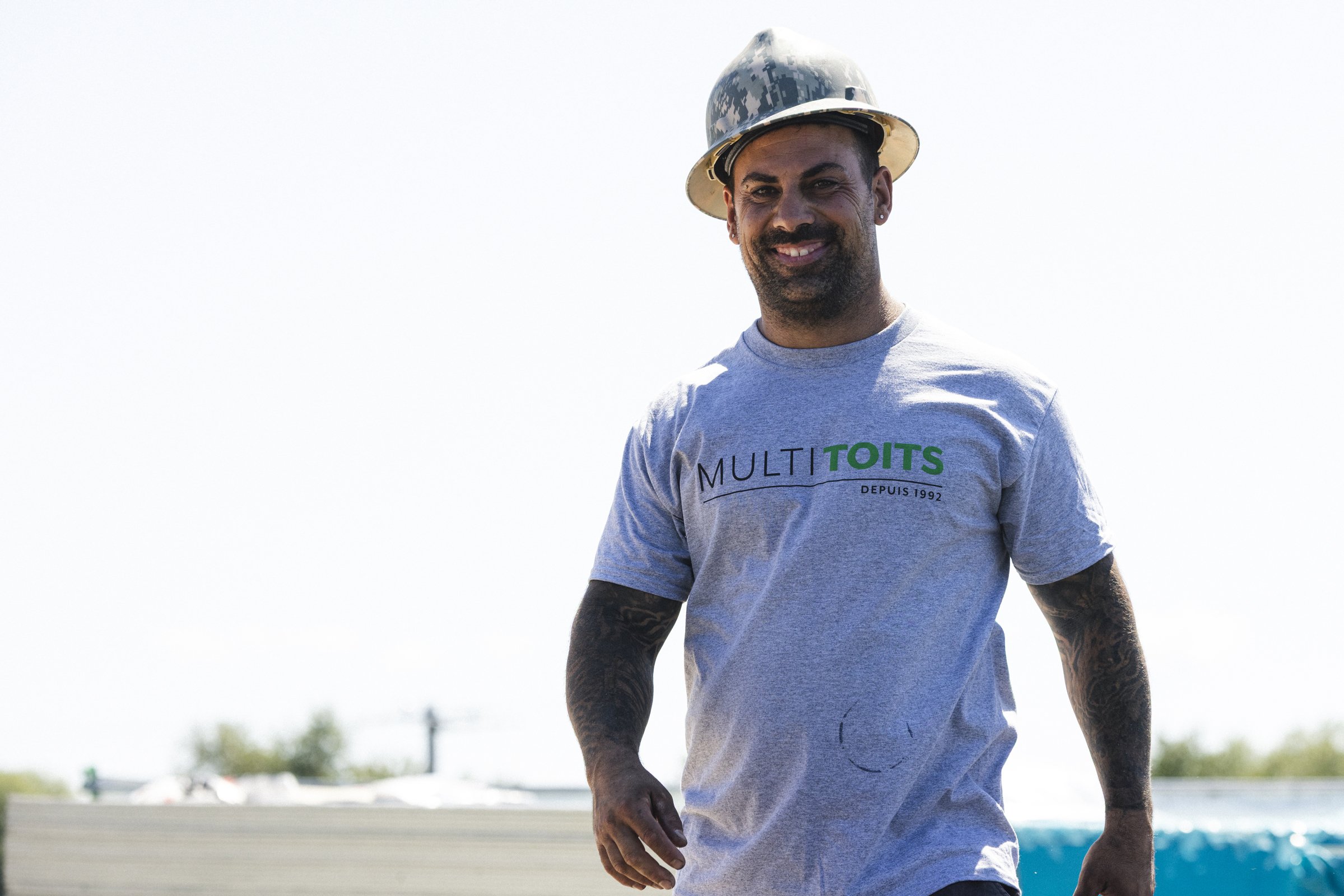 Homme portant un casque de sécurité et un t-shirt gris avec le texte 'MULTI TOITS' en gras, souriant, à l'extérieur.