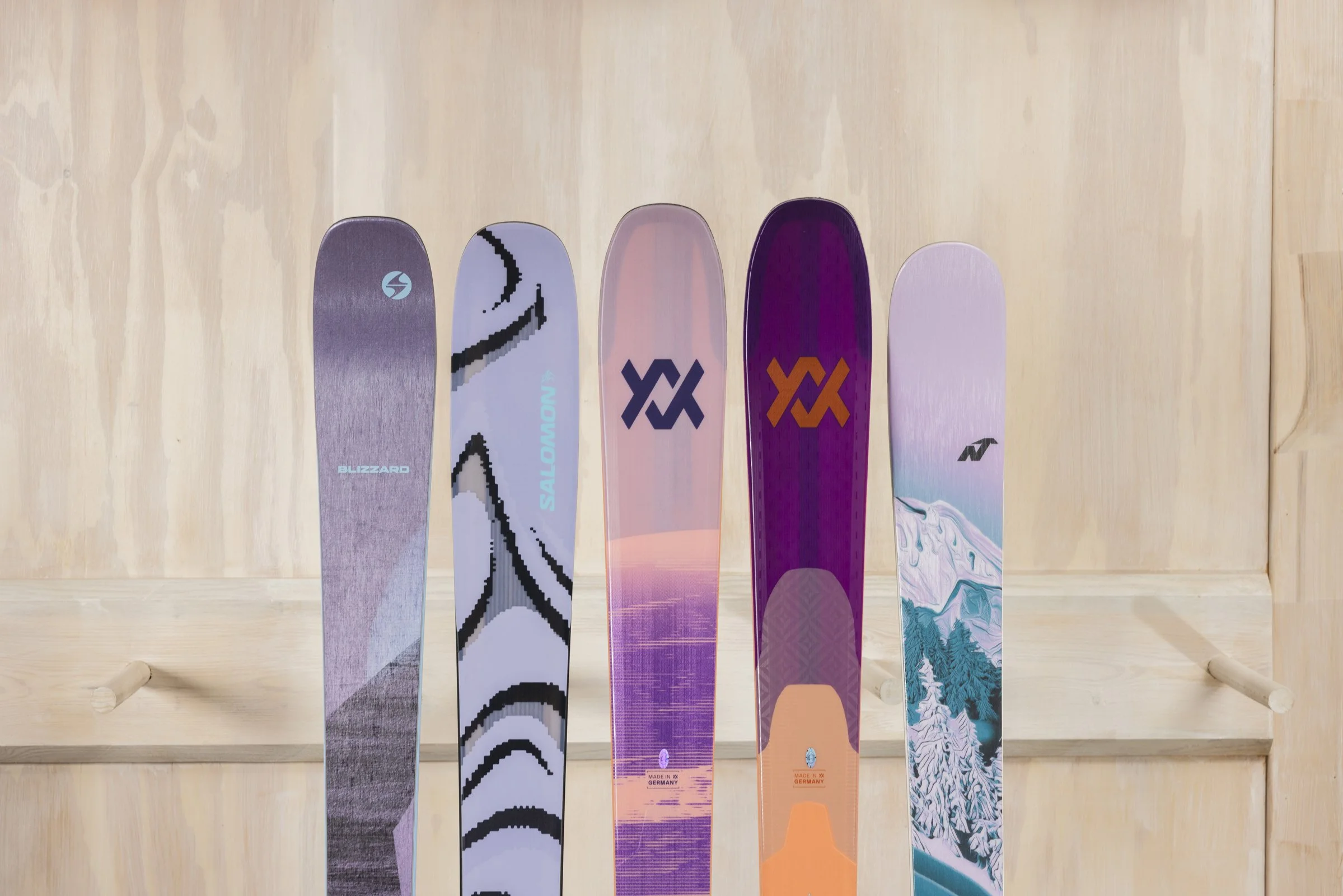 Cinq skis en ligne contre un mur en bois clair, chaque ski ayant un design différent et coloré.