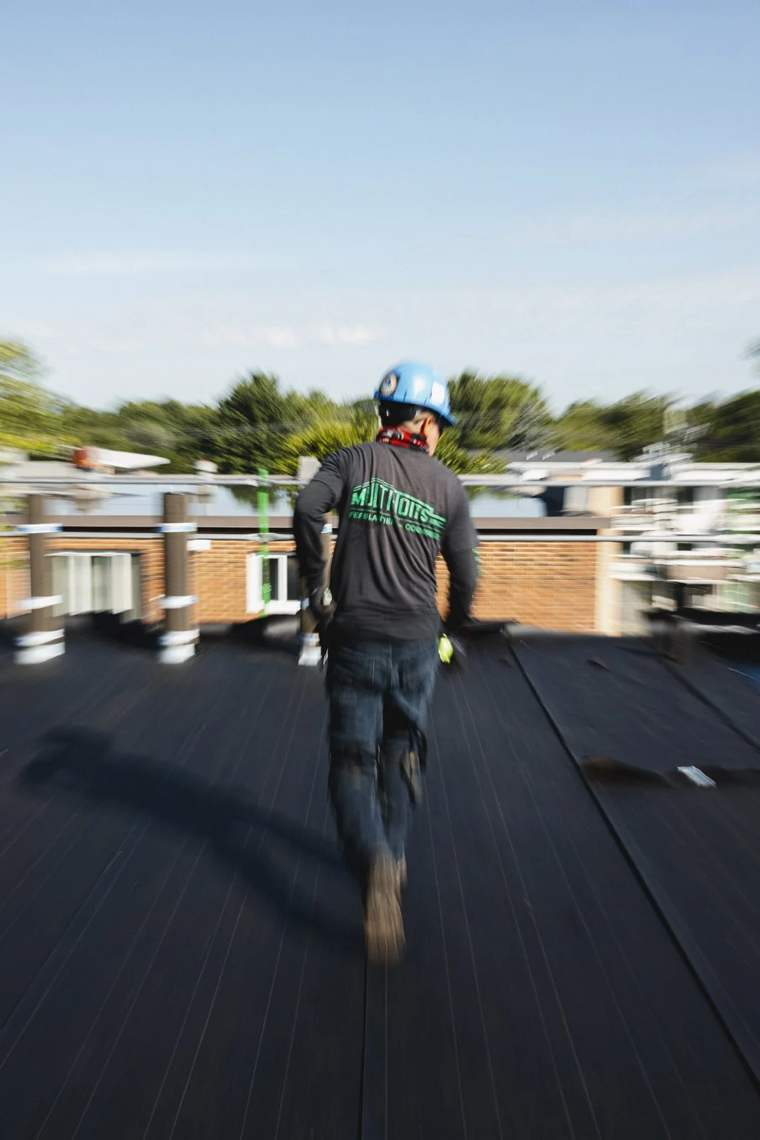 Un homme portant un casque bleu, un t-shirt noir avec un logo vert à l'arrière, marche sur un toit en construction. Arrière-plan flou d'immeubles et de ciel bleu.