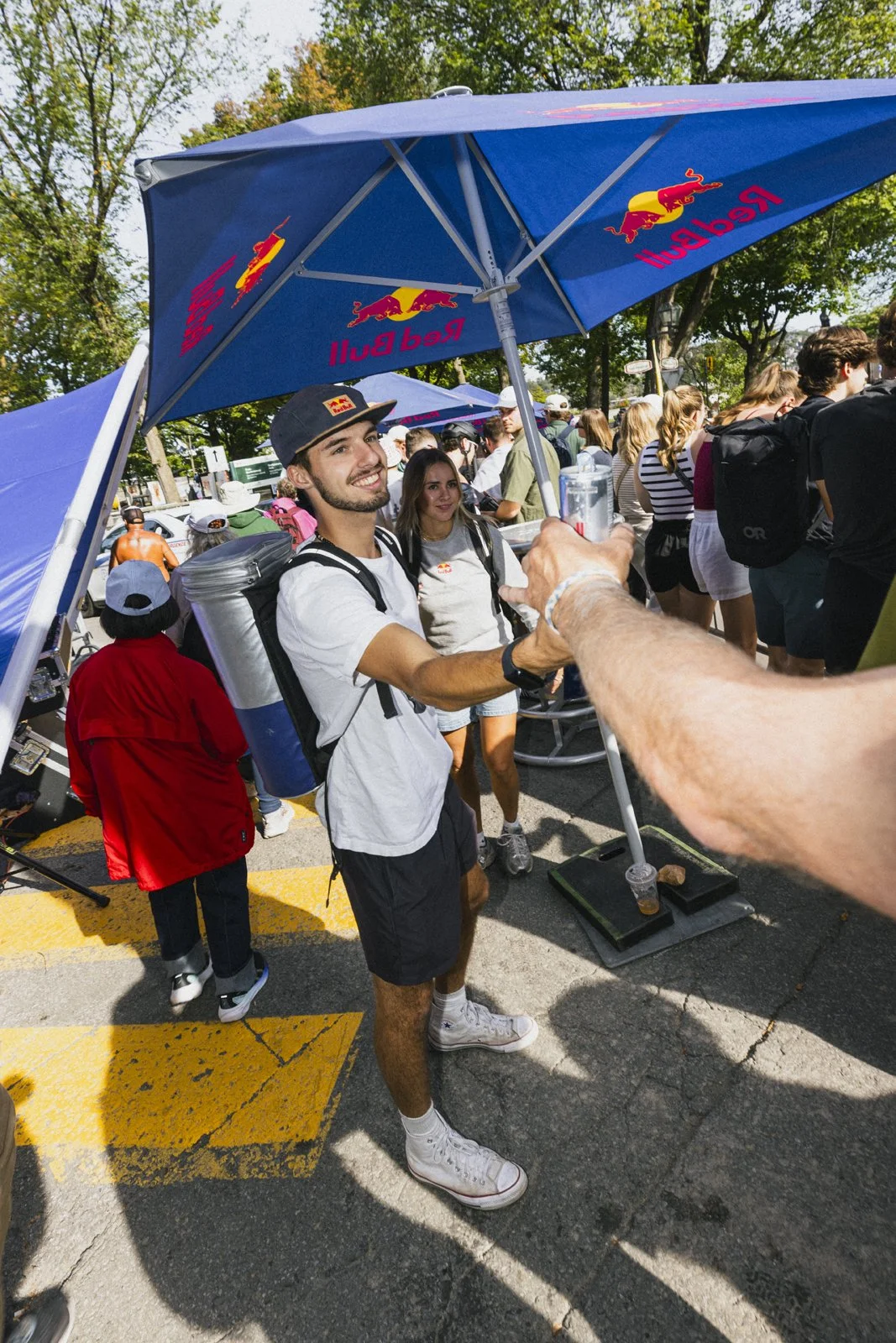 Un homme souriant, portant un sac à dos, donne une poignée de main à une personne non visible lors d'un événement en plein air avec de nombreuses personnes en arrière-plan. Un grand parasol Red Bull est visible.