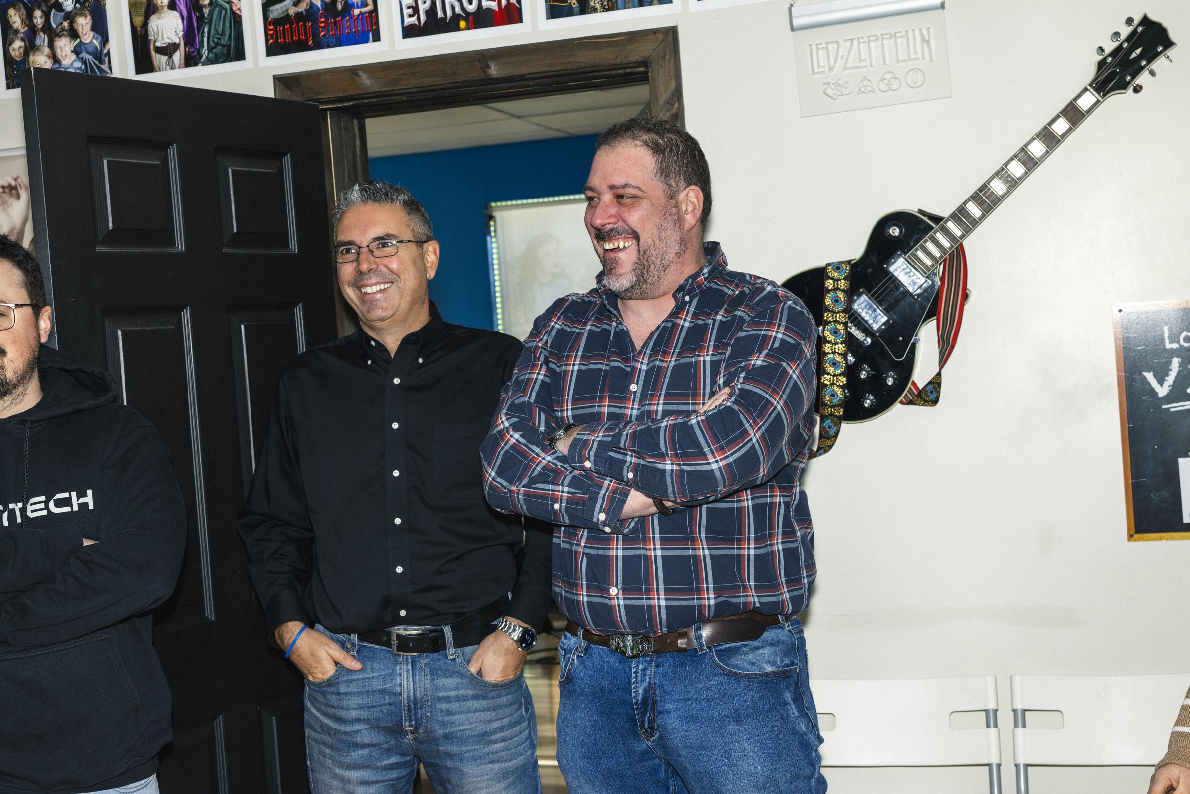 Trois hommes souriants, dont un avec les bras croisés, se tiennent dans une pièce décorée de posters de groupes de musique et d'une guitare électrique accrochée au mur.