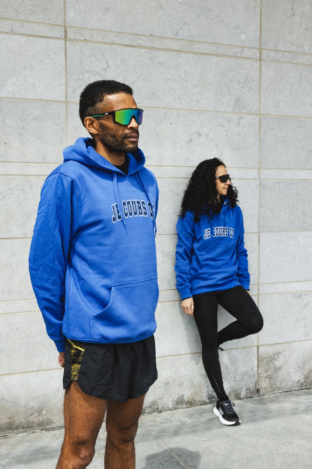 Deux personnes portant des sweatshirts bleus avec l'inscription "JL. COURS O" se tiennent contre un mur en pierre. L'homme devant porte des lunettes de sport à lunettes et un short noir, la femme derrière a des lunettes de soleil et une jambe levée.