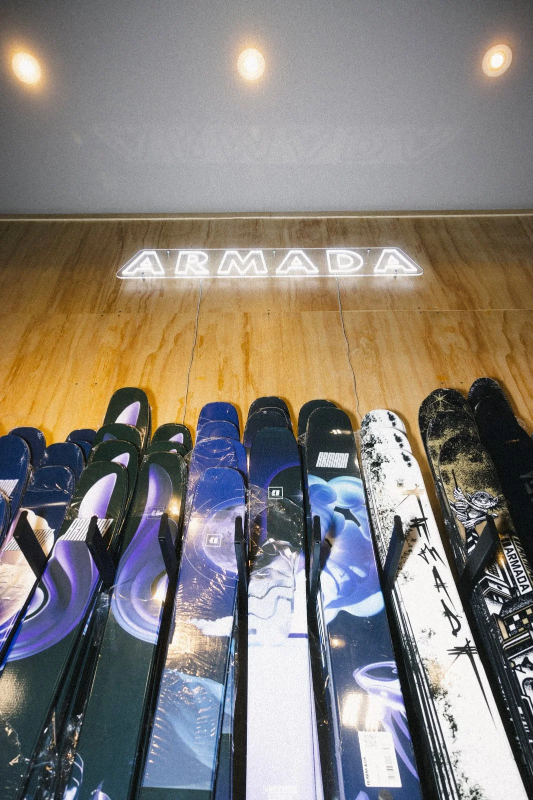 Skis de couleur noire, bleue, blanche et avec des motifs variés, alignés verticalement dans un magasin contre un mur en bois, avec une enseigne lumineuse au-dessus affichant le mot 'ARMADA'.