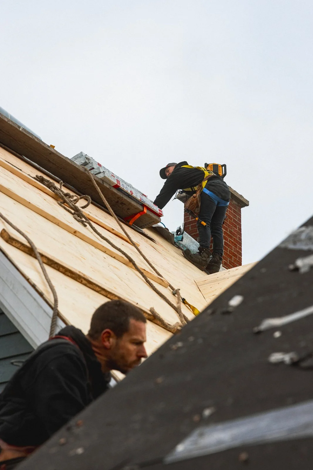 Deux ouvriers effectuent des travaux de roofing sur un toit en pente, l'un en haut à côté d'une cheminée en briques installée, l'autre en dessous, avec des outils à la main.
