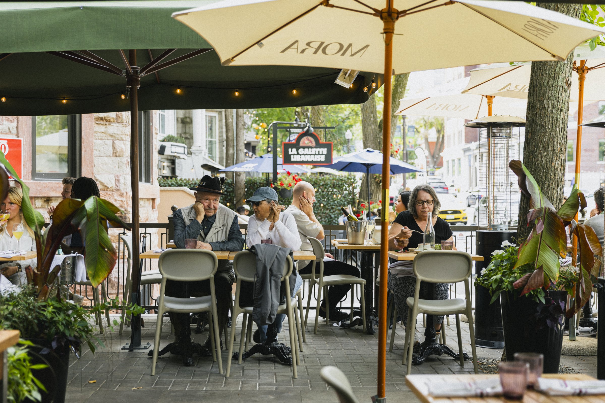 Terrasse de café en plein air avec plusieurs personnes assises à des tables, sous des parasols beige et verts. Certains clients dégustent, d'autres discutent. Des plantes décoratives entourent la terrasse et un bâtiment en pierre est visible en arriè