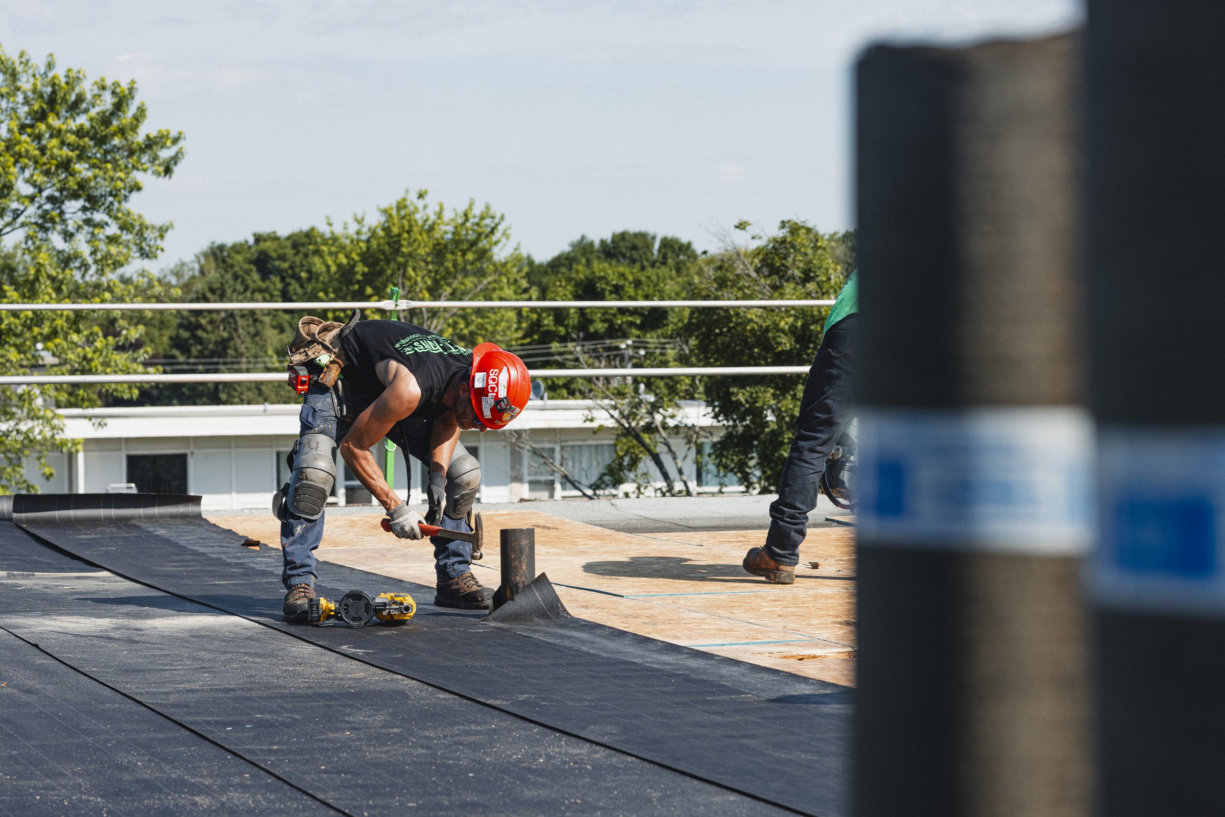 Ouvrier en équipement de sécurité travaillant sur le toit à l'extérieur, posant une membrane de toiture avec outils.