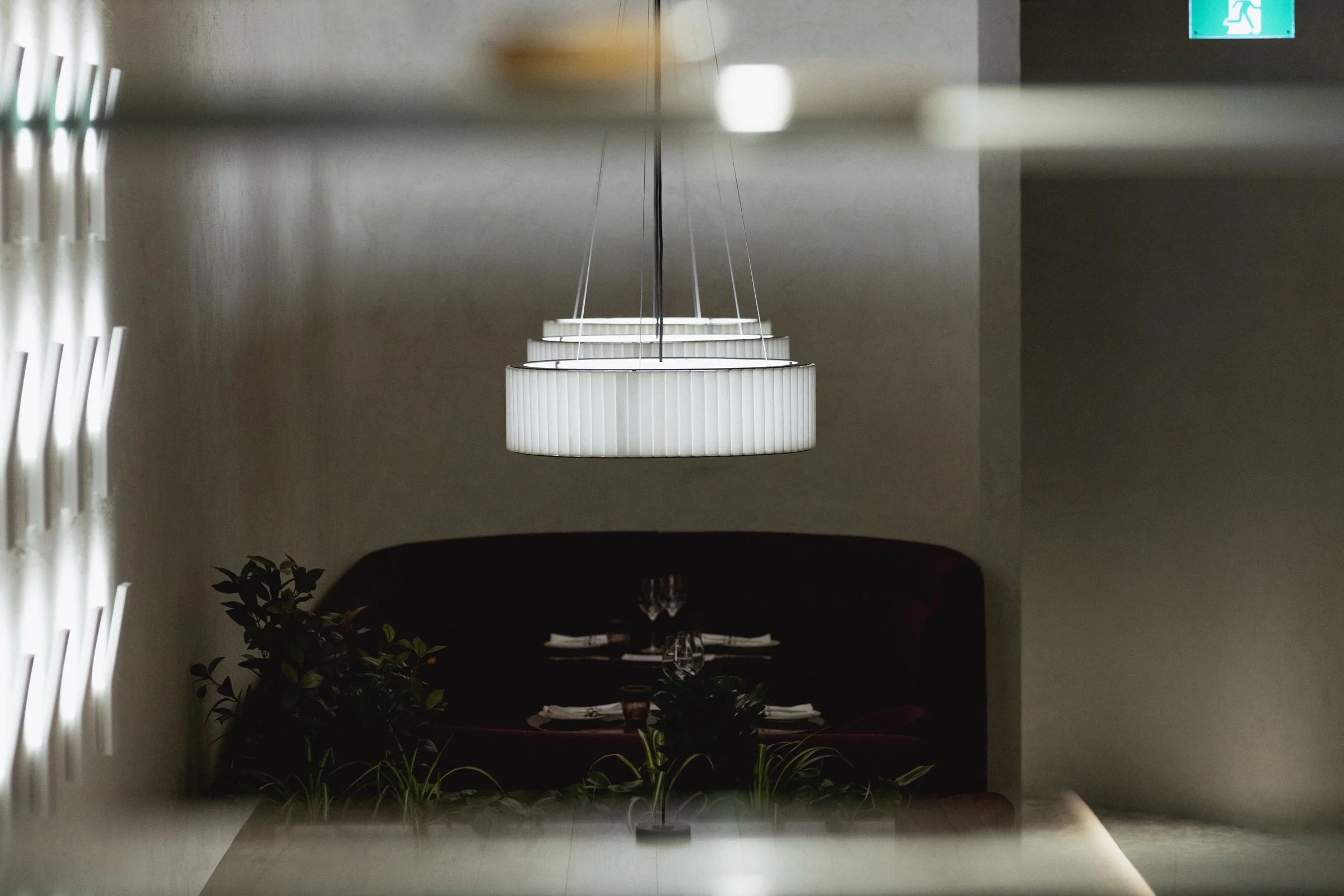 Une salle à manger avec un grand luminaire blanc suspendu au plafond. Il y a une table prête pour un repas, entourée d'une banquette sombre et décorée de plantes vertes. Un panneau de sortie de secours est visible sur le mur en arrière-plan.