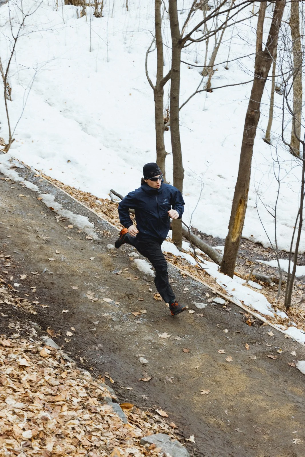 Un homme court sur un sentier de forêt en hiver, avec des feuilles mortes et de la neige autour.