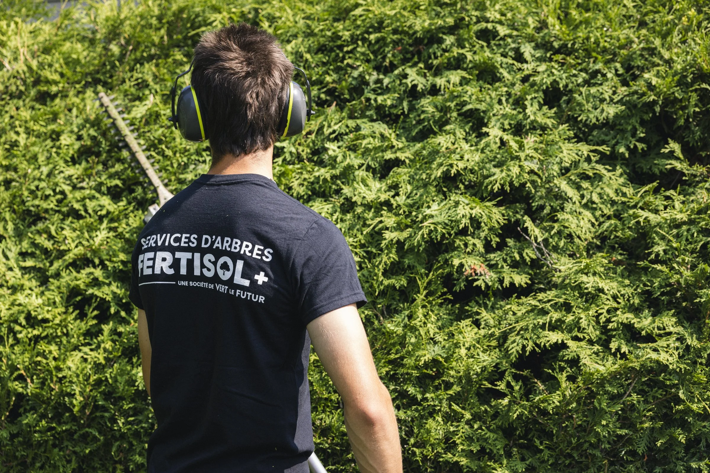 Un jeune homme portant un t-shirt noir avec l'inscription 'Services d'Arbres FERTISOL' stylisée, en train de couper ou d'entretenir des arbustes dans un espace extérieur avec des arbustes verts.