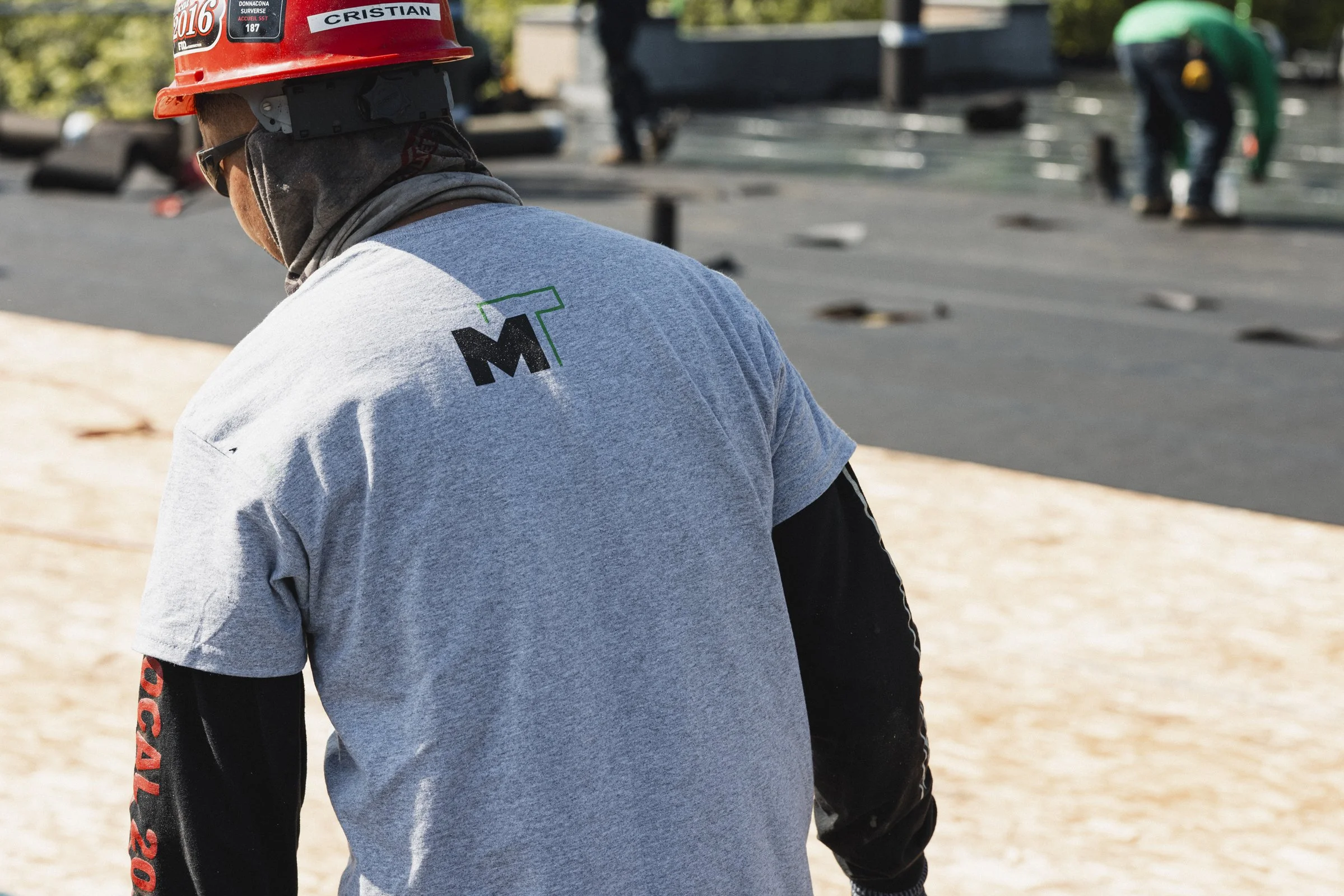 Travailleur portant un casque rouge avec une étiquette à son nom, travaillant à l'extérieur sur un chantier ou une construction.