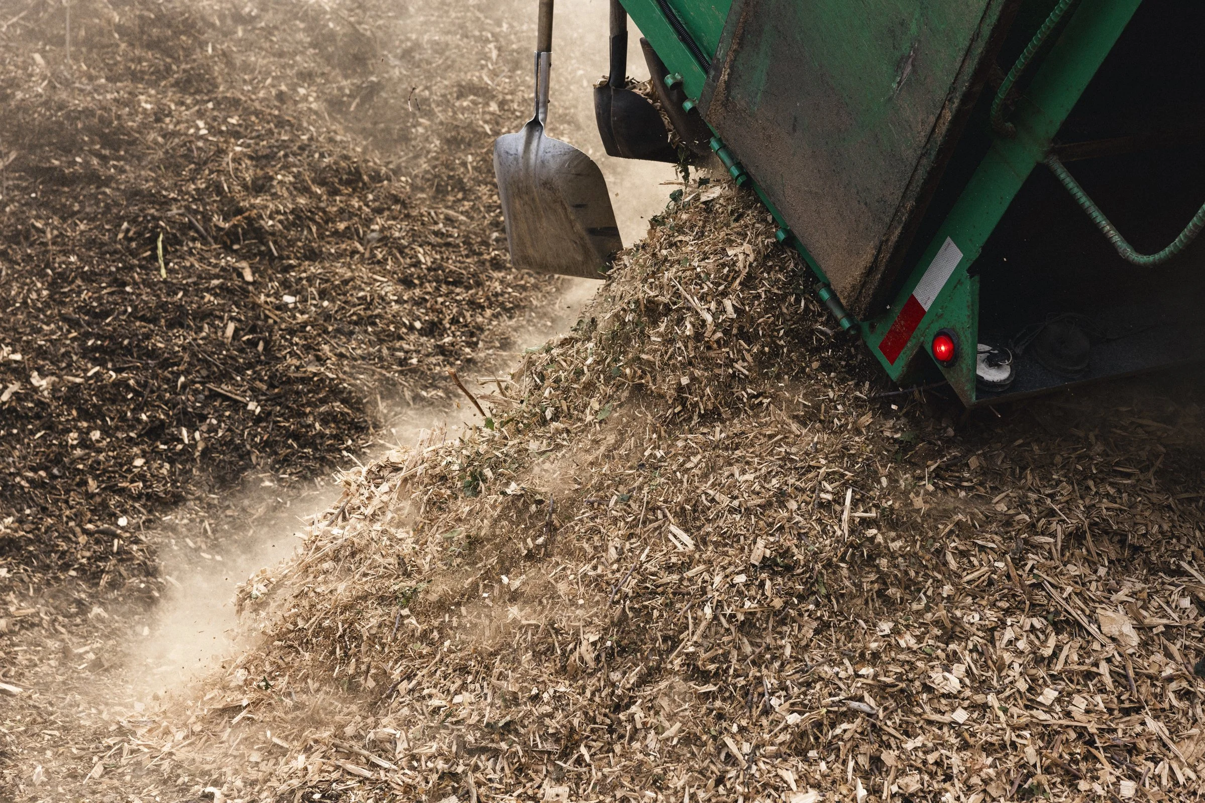 Une machine de traitement de compost transformant des déchets de bois en compost, avec une pelle en retrait et de la poussière en suspension.