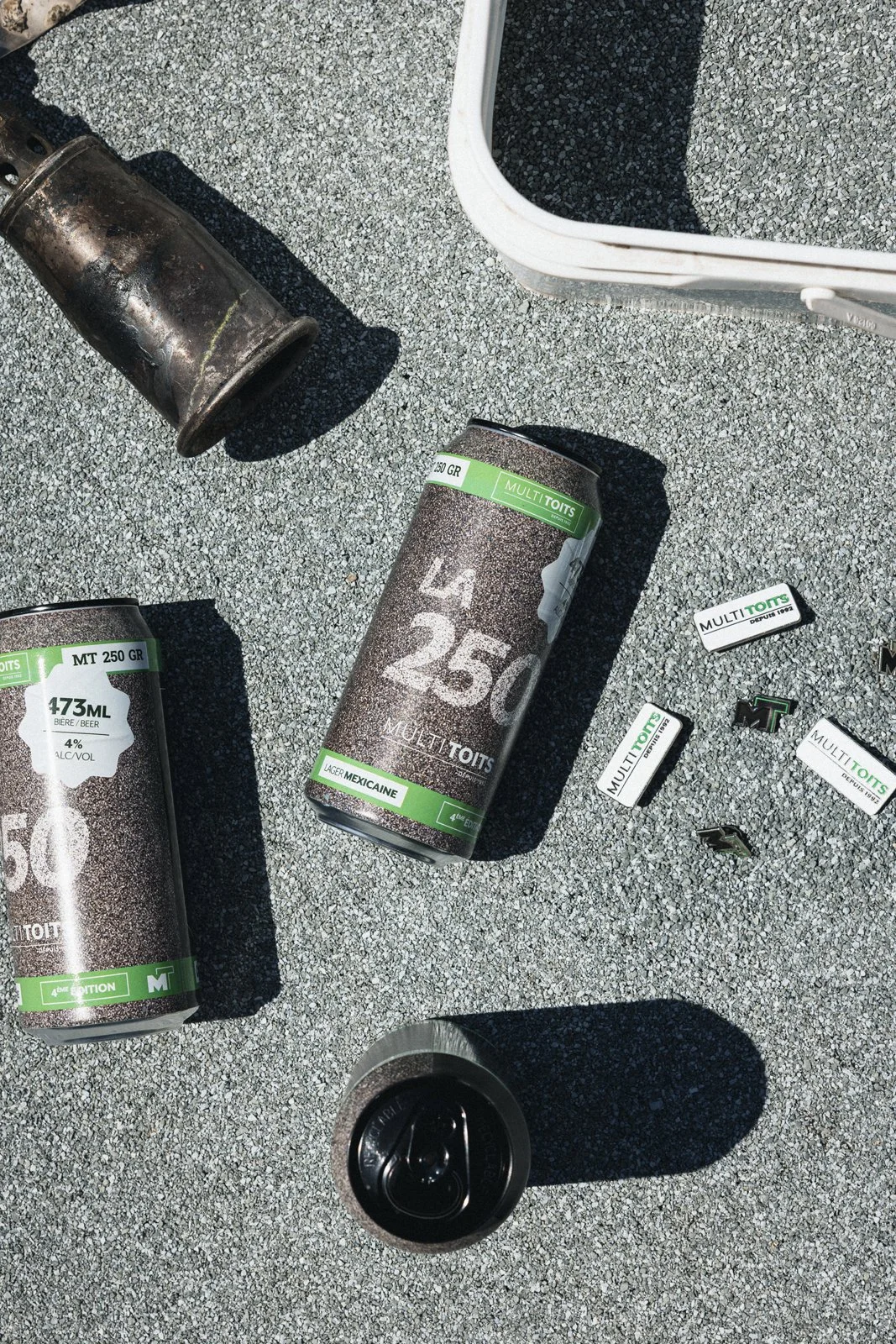Des canettes de bière, des bouchons et des morceaux de papier avec le logo 'MULTITOITS' sur une surface grise, avec un seau blanc et une tasse en métal.