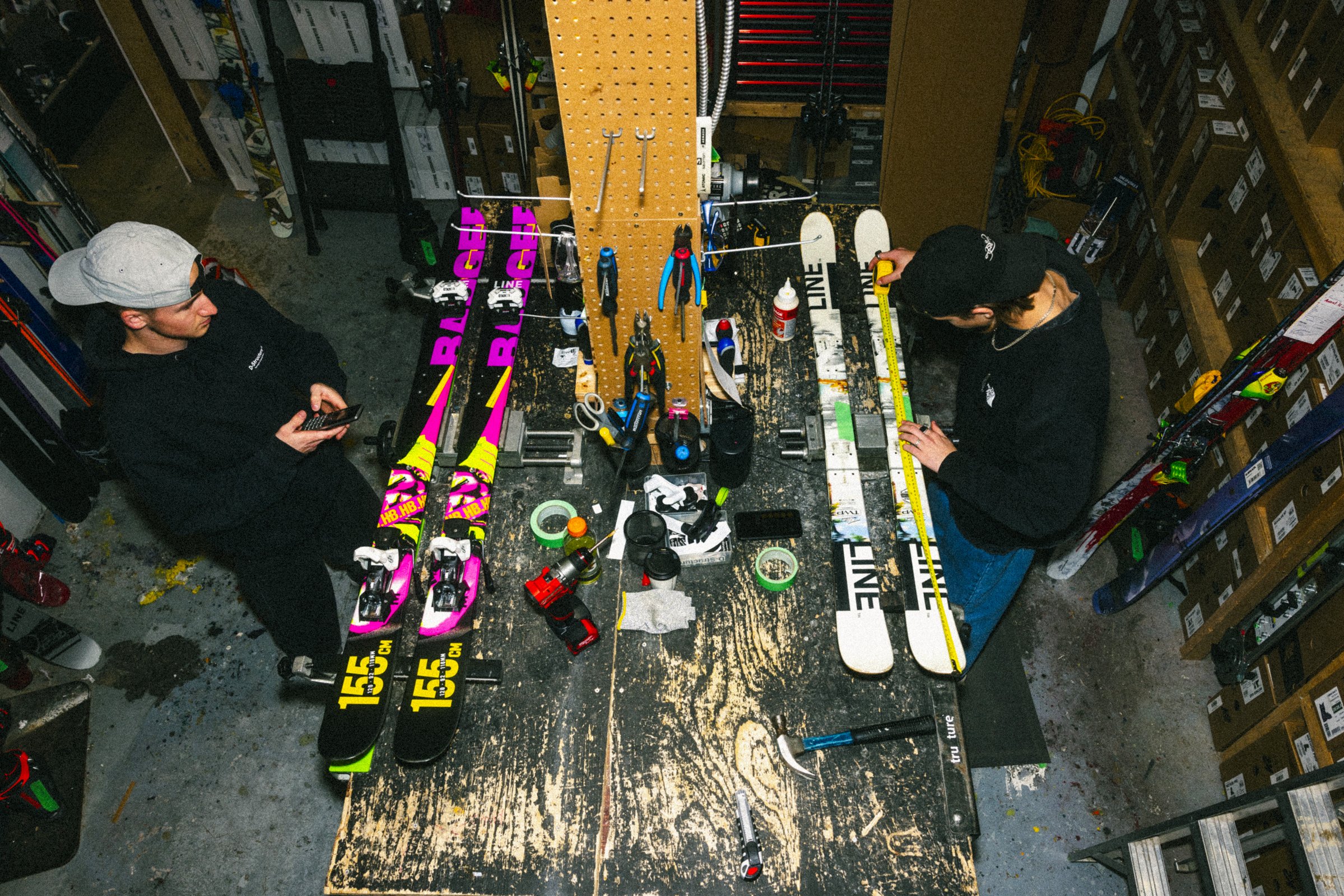 Deux personnes en train d'assembler des skis dans un atelier. Le plan est vu du dessus et montre une table de travail chargée d'outils, de rubans adhésifs, de tournevis, de colle et d'autres équipements. Un des individus porte une casquette blanche, 