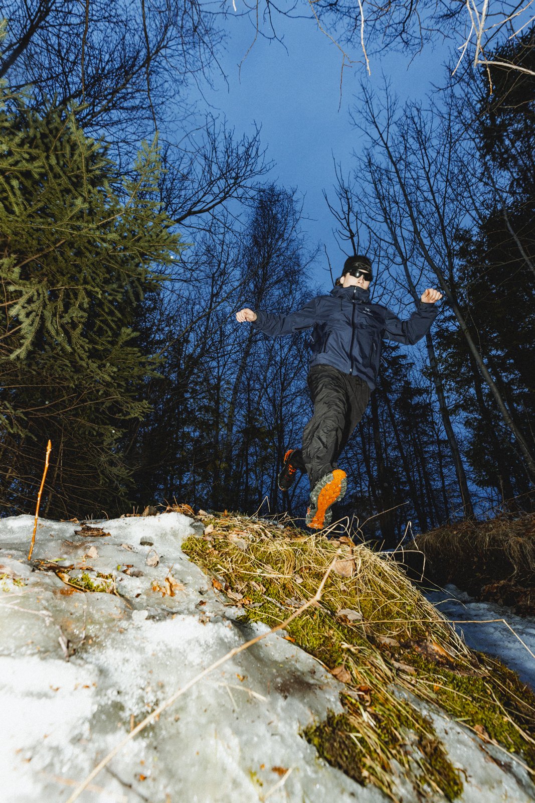 Un homme en vêtements de randonnée, portant des lunettes de soleil, court ou descend une pente recouverte de glace et de neige dans une forêt en hiver, au crépuscule.