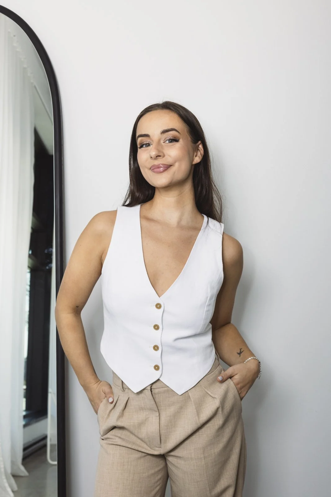 Jeune femme souriante avec des cheveux bruns, portant un haut blanc à boutons et un pantalon beige, posant contre un mur blanc, avec un miroir à gauche.