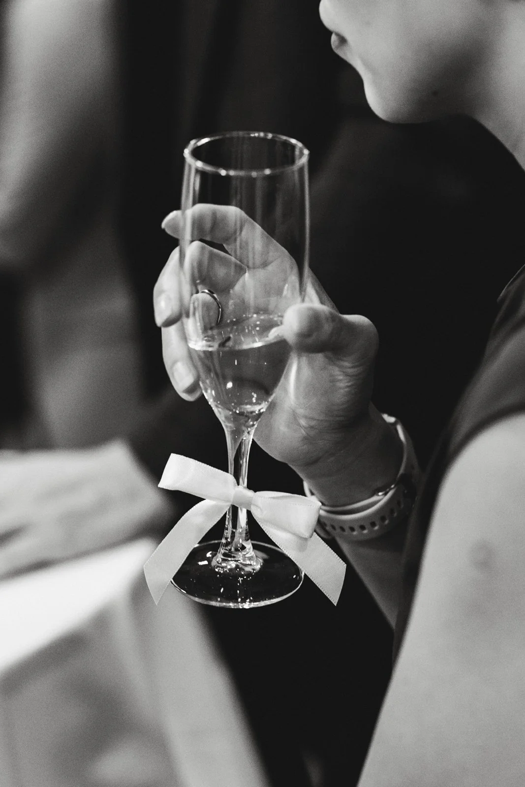 Main d'une personne tenant une flûte à champagne avec un ruban blanc attaché au pied, la personne porte une montre et un anneau, et semble être en contexte de célébration ou d'événement spécial.