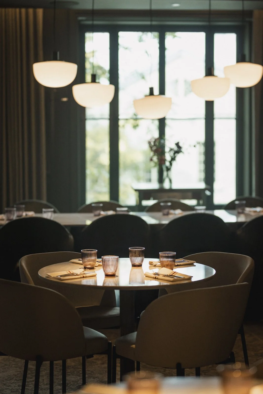 Une salle à manger élégante avec une table ronde dressée avec des verres et des couverts en face de grandes fenêtres laissant entrer la lumière naturelle. Des lampes suspendues blanc cassé suspendues au plafond.