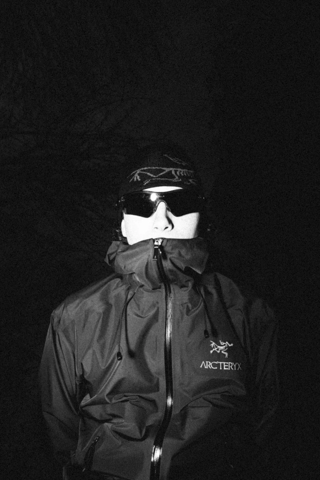 Personne portant une veste de randonnée, lunettes de soleil et un bandeau, photo en noir et blanc, dans un environnement sombre.