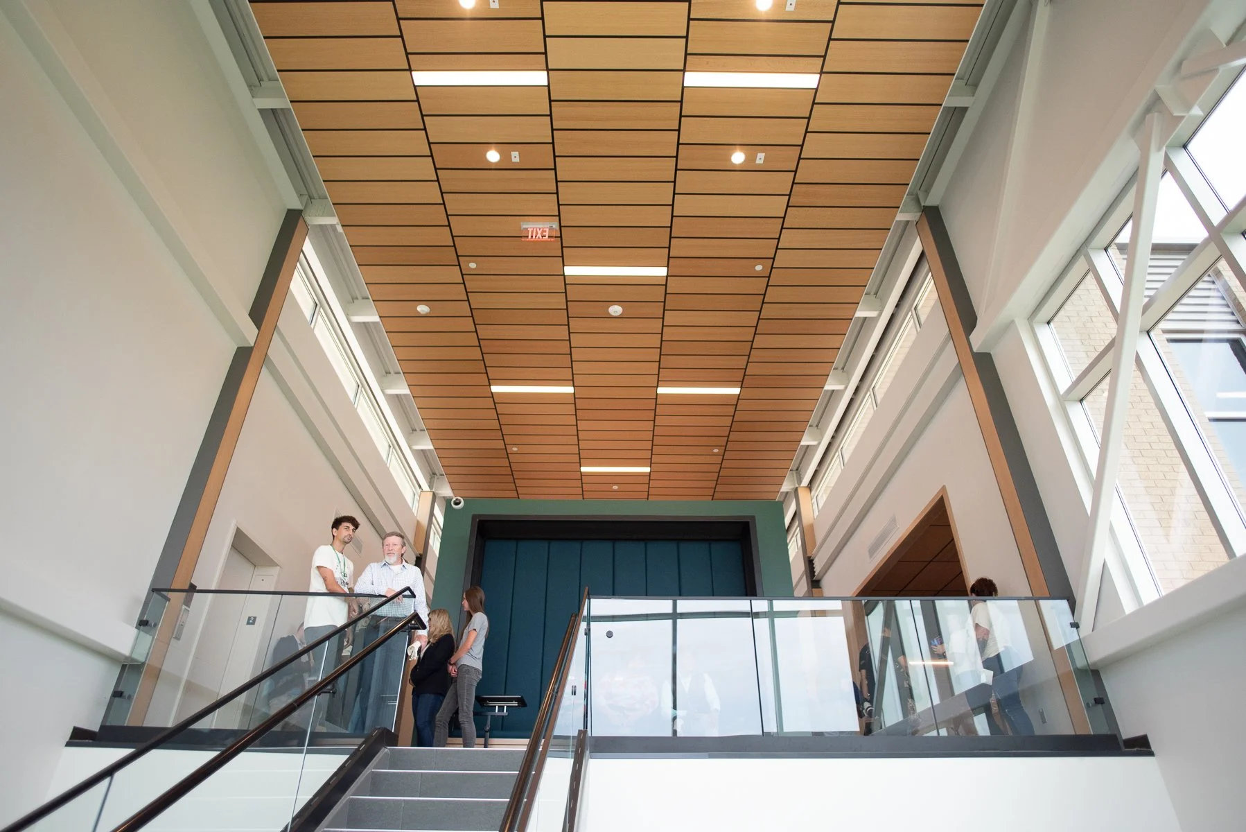 Large windows illuminate the blonde wood ceiling at the top of a modern staircase while people stand and chat.