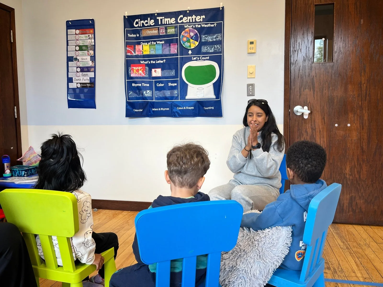 A young woman is sitting in front of three children, engaging them in a group activity inside a classroom. The children are seated on colorful plastic chairs and are listening attentively. Behind them, there is a blue educational bulletin board titled 'Circle Time Center' with various charts and educational content.
