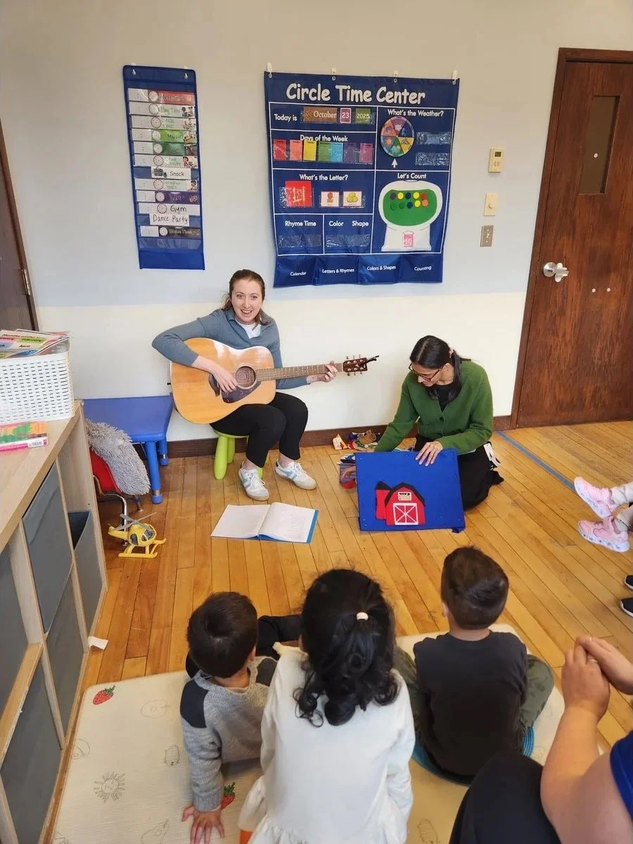 A woman is playing guitar and singing to children seated on the floor in a classroom. Another woman is sitting nearby, holding a picture book. The classroom has educational posters on the walls and a small rug with children’s drawings.