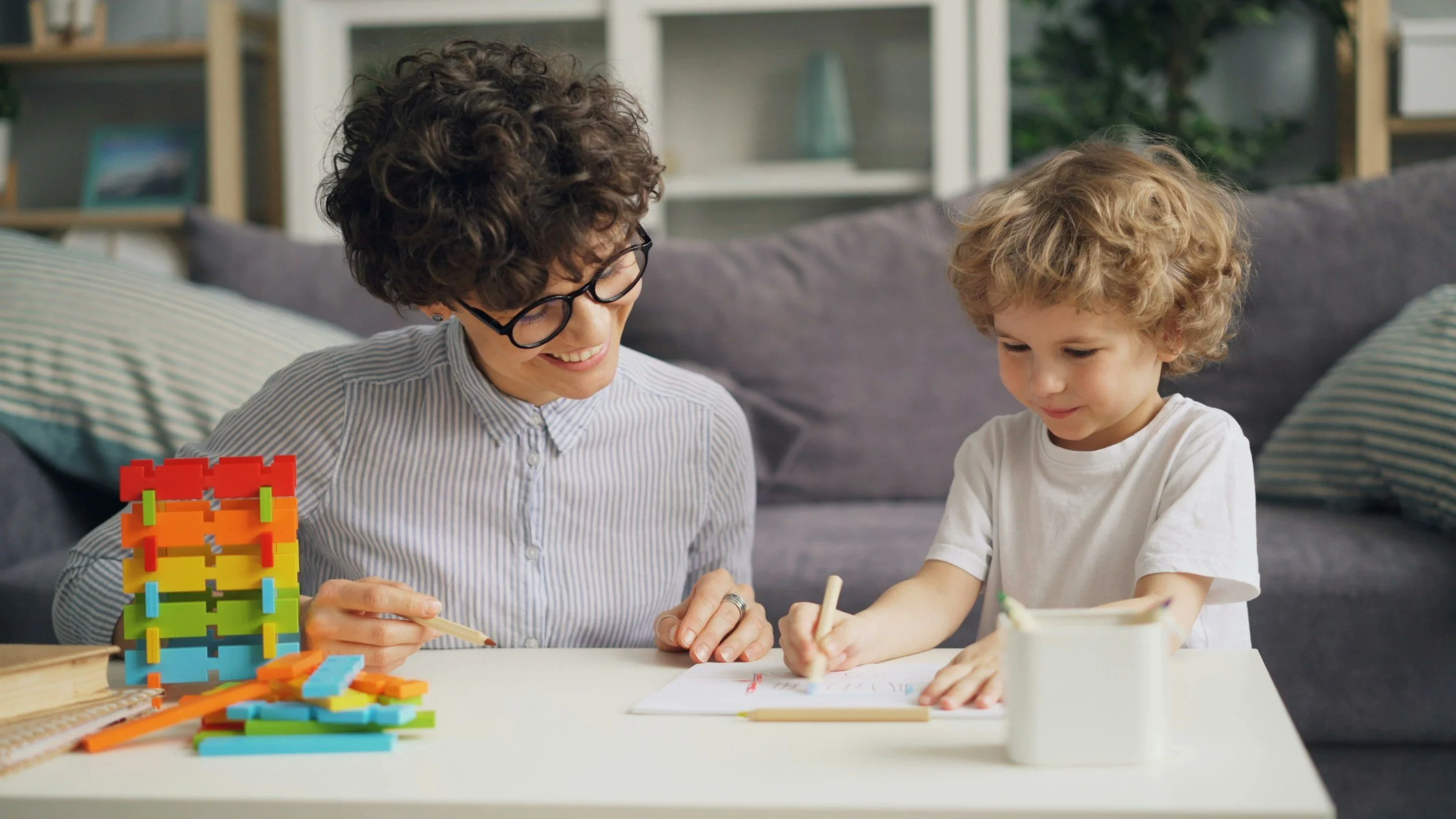 Adult woman and young boy drawing with colored markers at a white table, with colorful building blocks and a white container nearby, in a cozy living room.