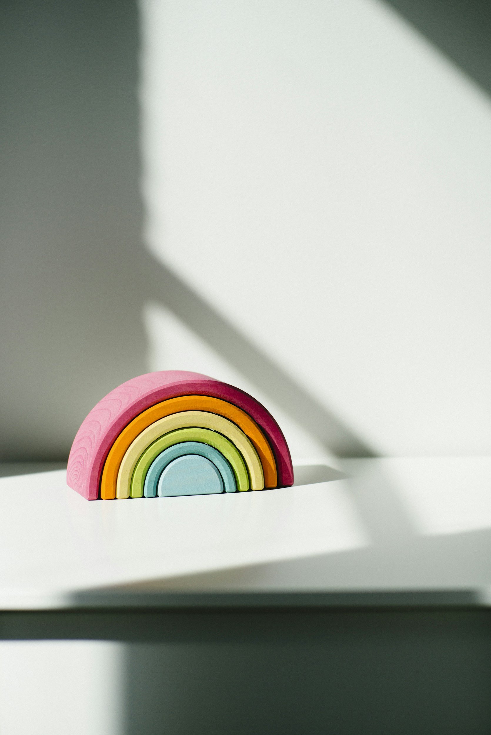 Colorful wooden rainbow toy on a white surface with shadows and light patterns.