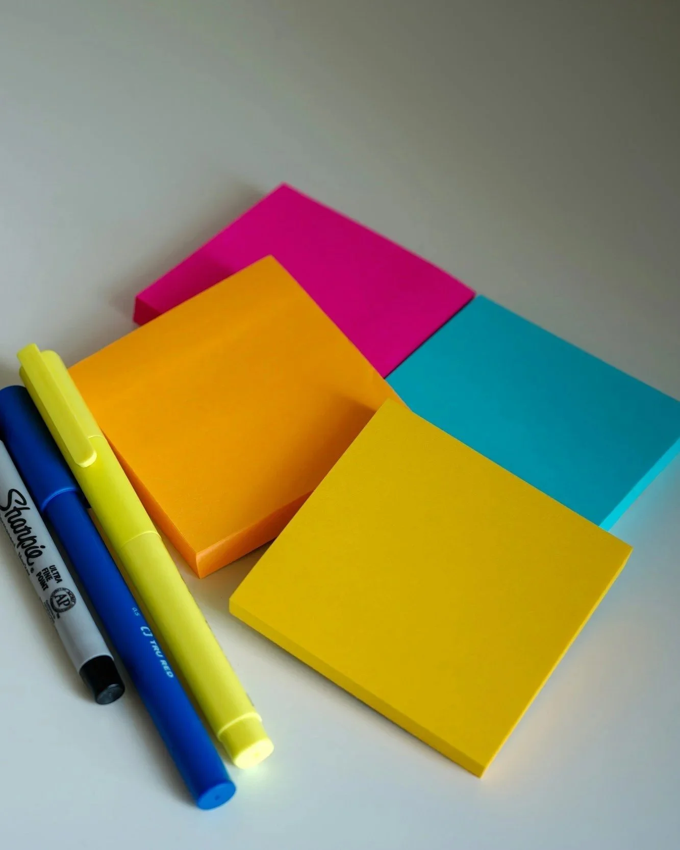 A collection of colorful sticky notes in pink, blue, yellow, and orange, with blue, yellow, and black markers on a white surface.