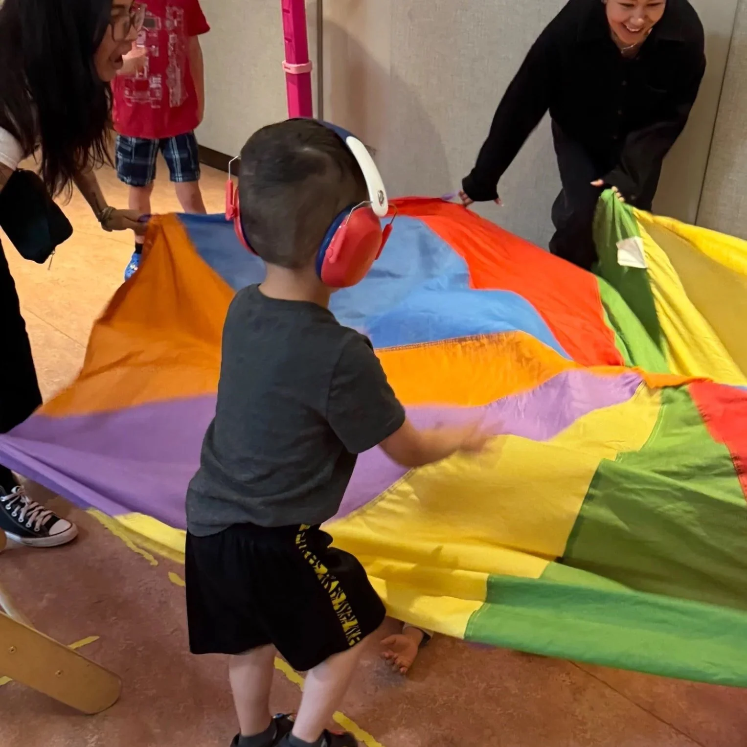 Children and adults playing with a colorful parachute indoors, with one child wearing headphones.