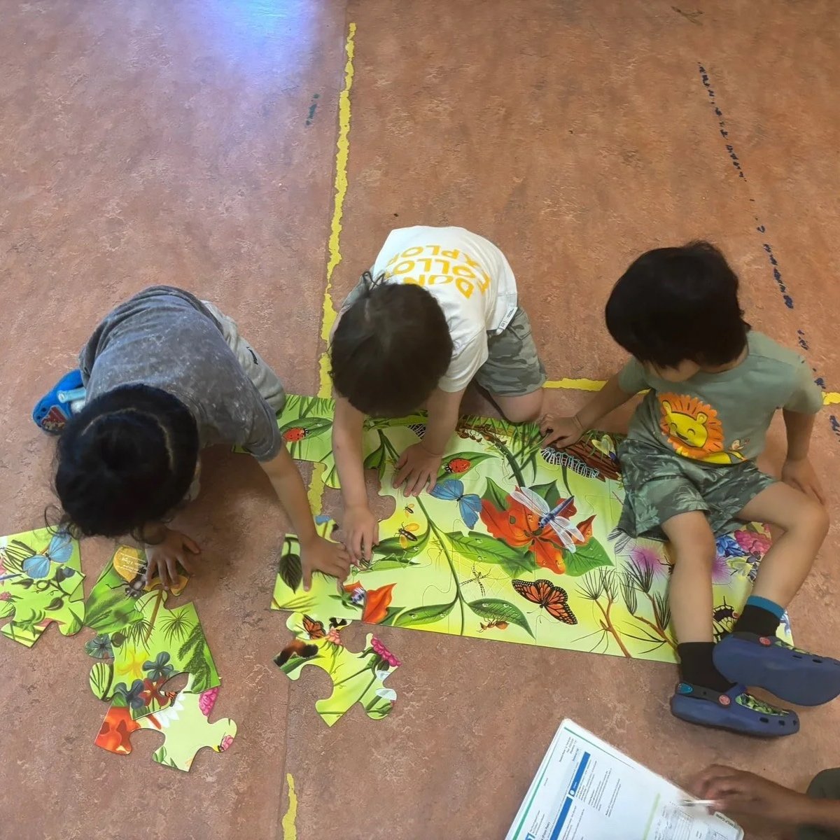 Three children working on a colorful butterfly-themed jigsaw puzzle on the floor of an indoor space.