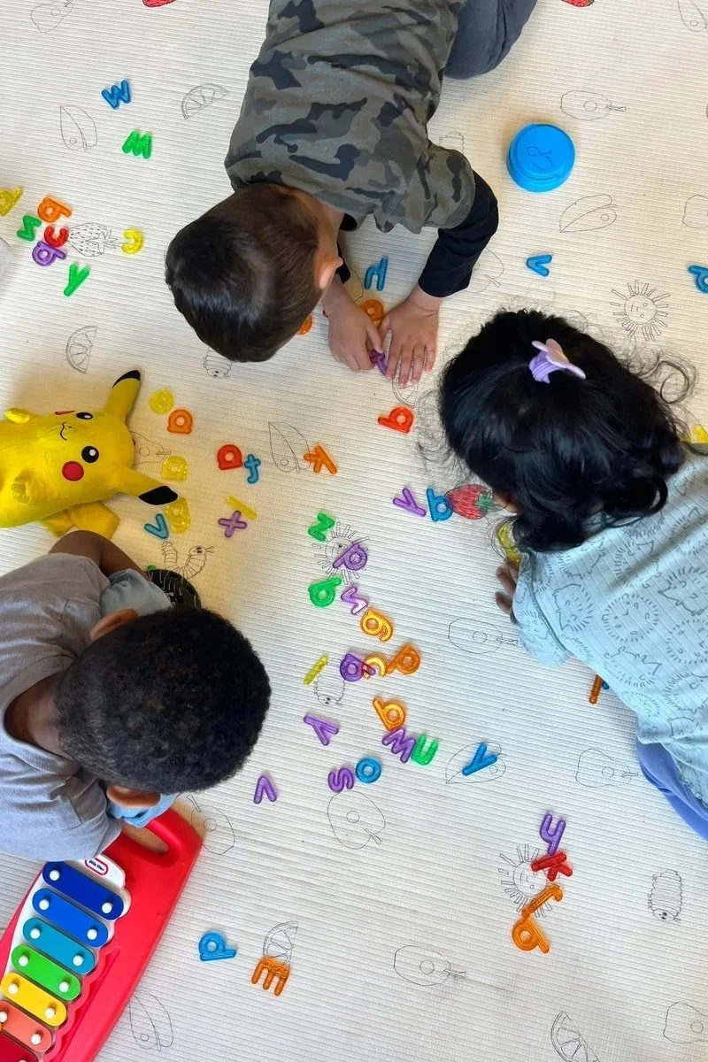 Three children playing with colorful magnetic letters on a white surface. A Pikachu plush toy is nearby, and a red toy xylophone is partially visible. The surface has light sketches of animals and suns.