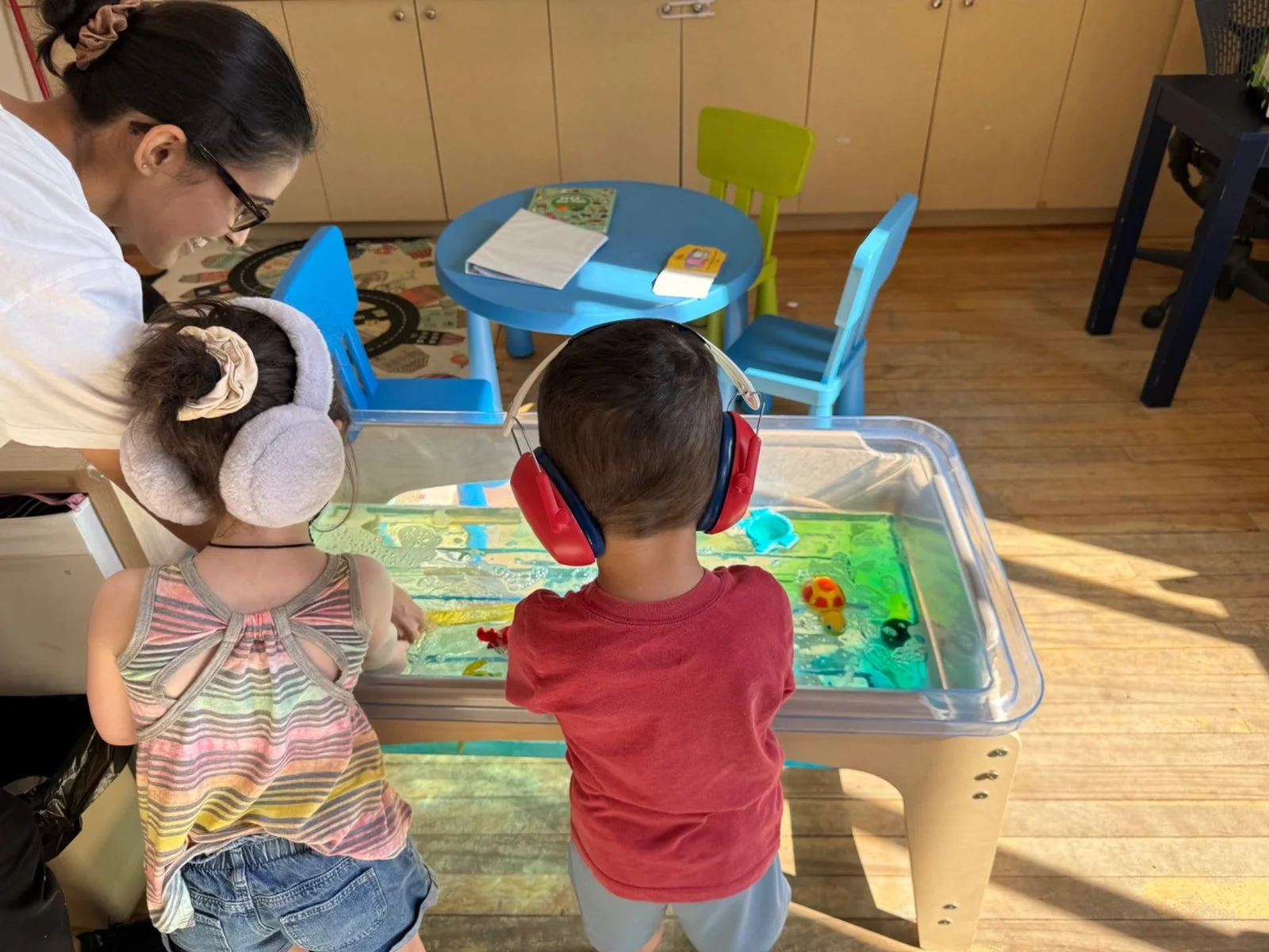 Two children playing at a water table with a woman supervising them in a classroom. One girl is wearing earmuffs and the other boy is using red headphones.