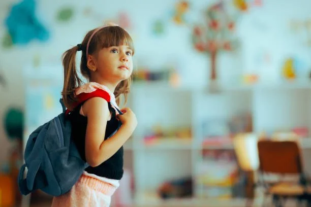 A young girl with pigtails carrying a backpack in a colorful classroom.