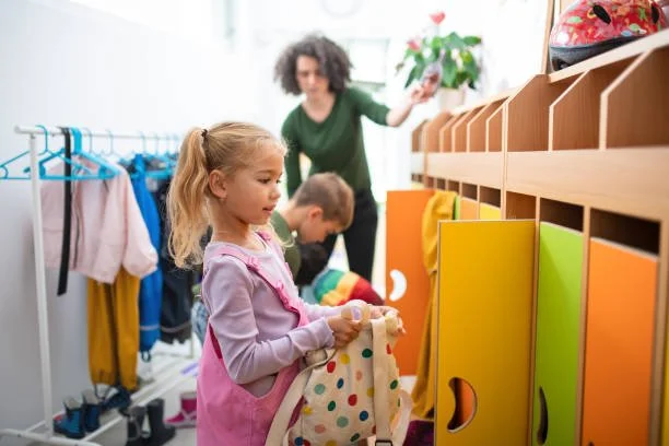 Young girl organizing a bag in a classroom with shelves, a teacher reaching for a plant, and a boy in the background.