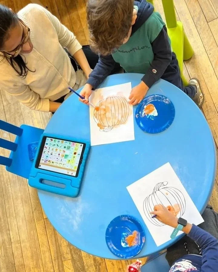 Two children and an adult decorating pumpkin outlines with orange paint at a blue round table, with a tablet displaying emojis nearby.