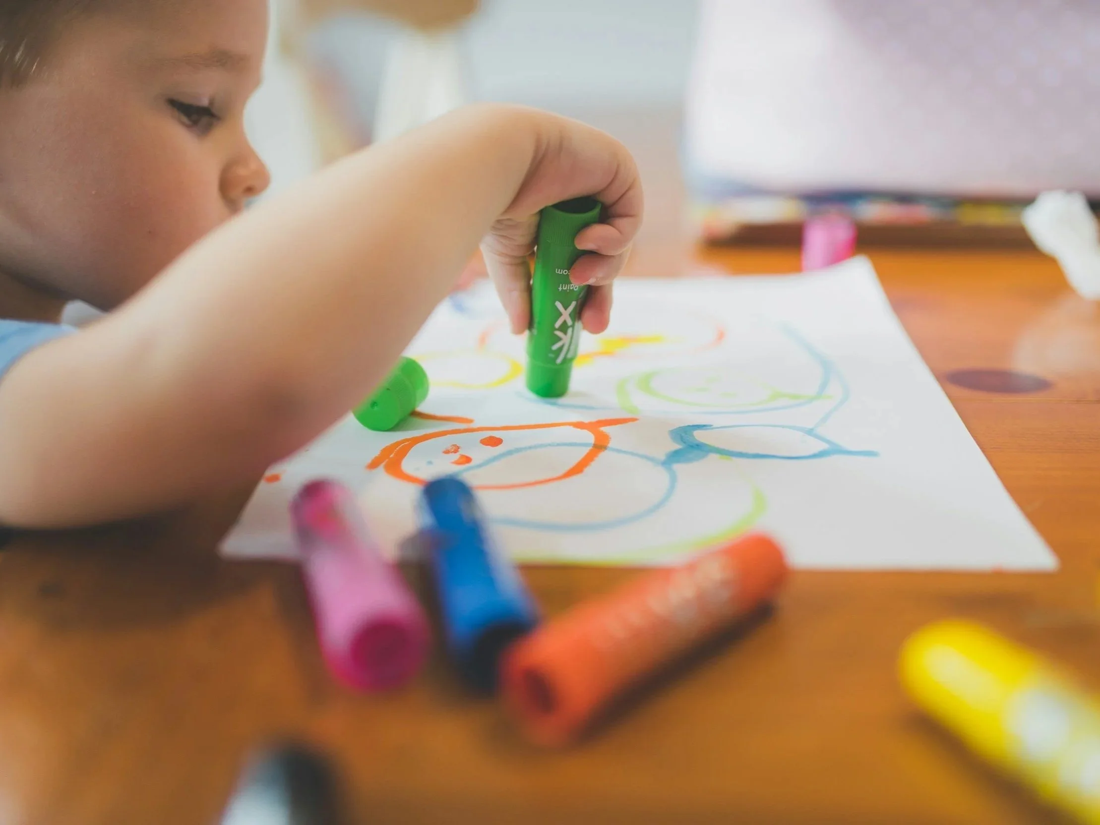 Child drawing on paper with colorful markers, focusing on their hand and face in profile, with scattered markers on a wooden table.