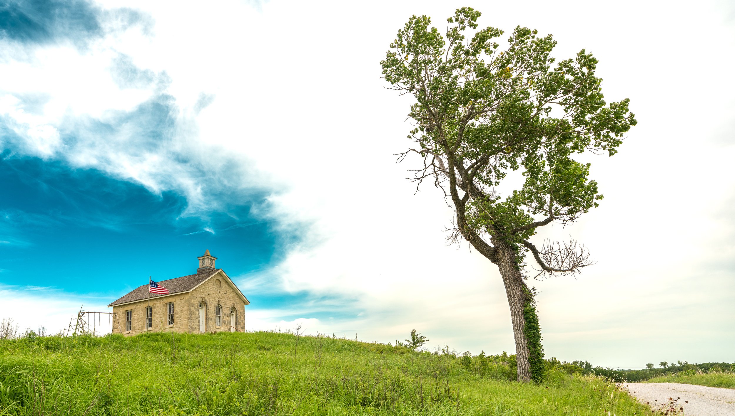 A large green tree standing beside a stone house with a watchtower and American flag, under a partly cloudy sky with a bright horizon.