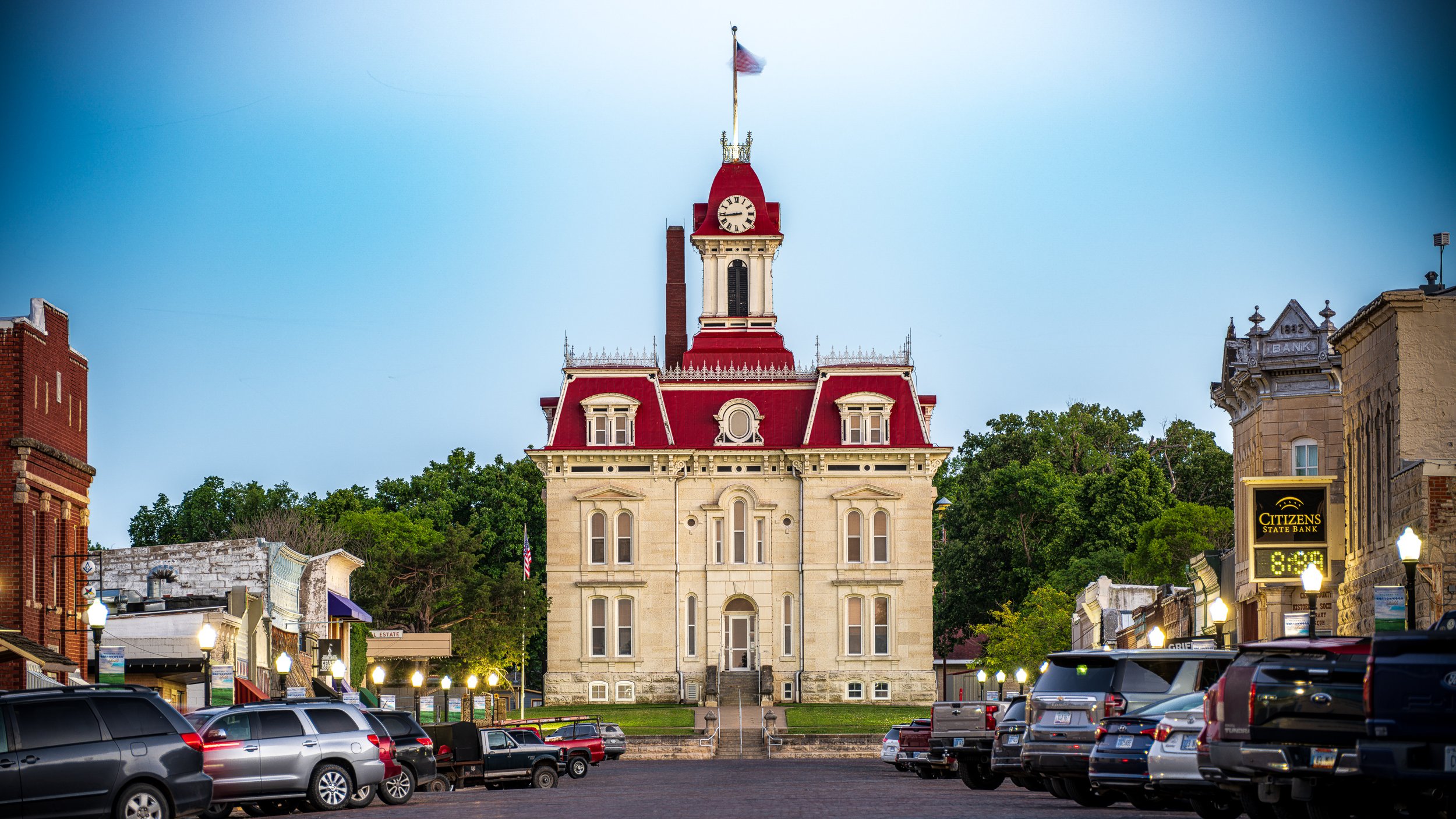 A historic courthouse with a clock tower and American flag, surrounded by parked cars and small storefronts in a downtown area.