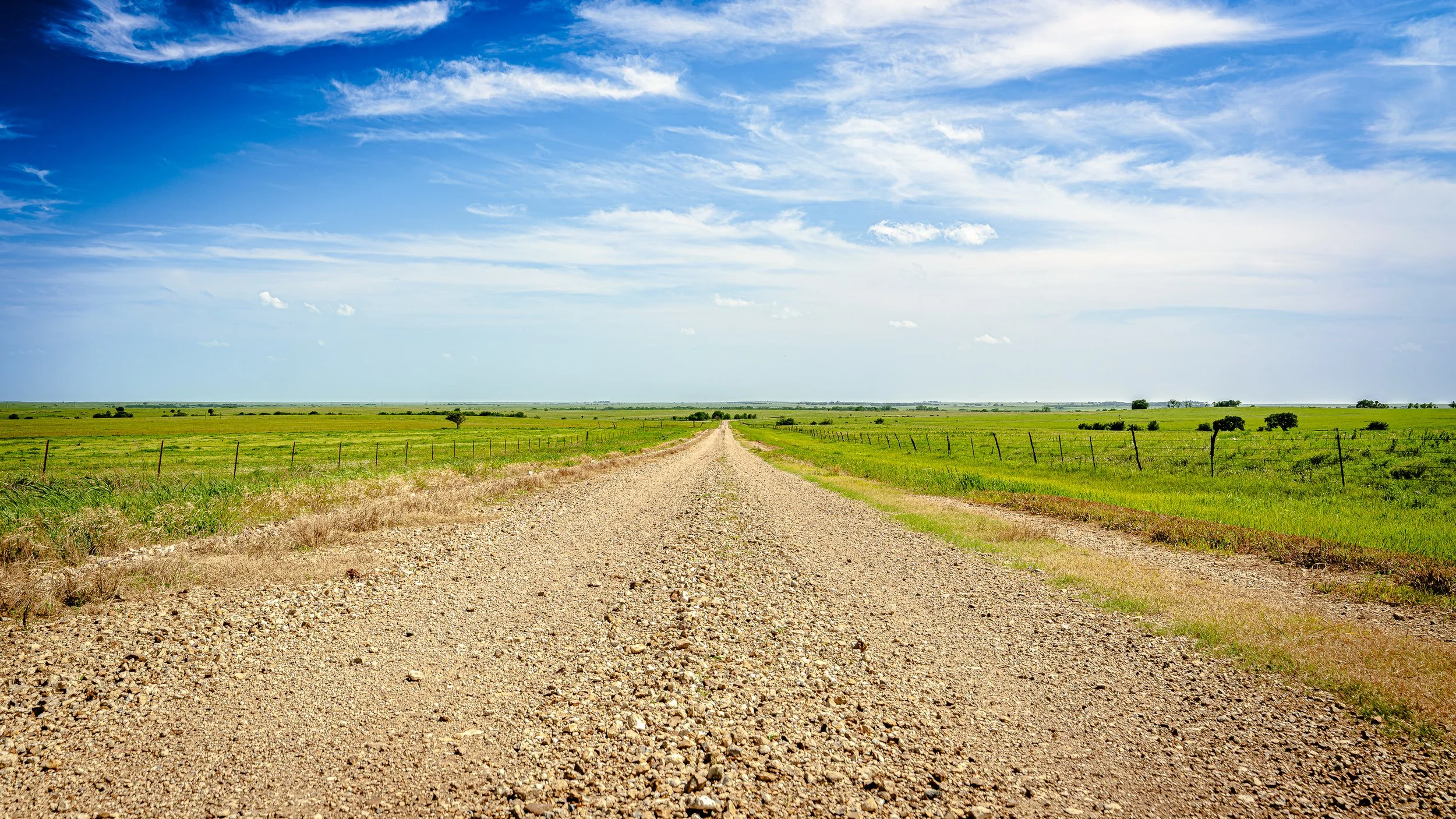 A gravel country road cutting through green fields under a blue sky with scattered white clouds.