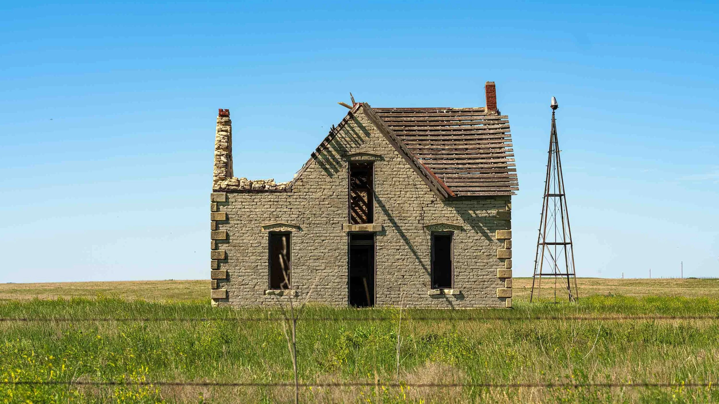 An old, abandoned brick house with a missing roof and broken windows, situated on a grassy field with a windmill nearby, under a clear blue sky.