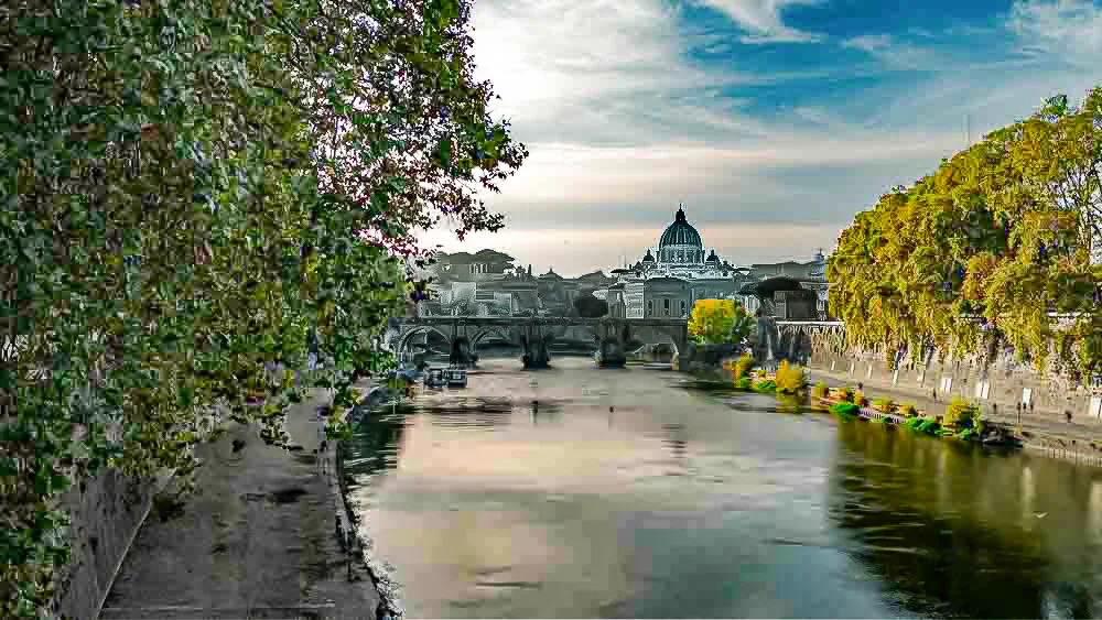 A scenic view of the Tiber River in Rome with a historic bridge, trees on both sides, and the Vatican's St. Peter's Basilica dome in the background under a partly cloudy sky.