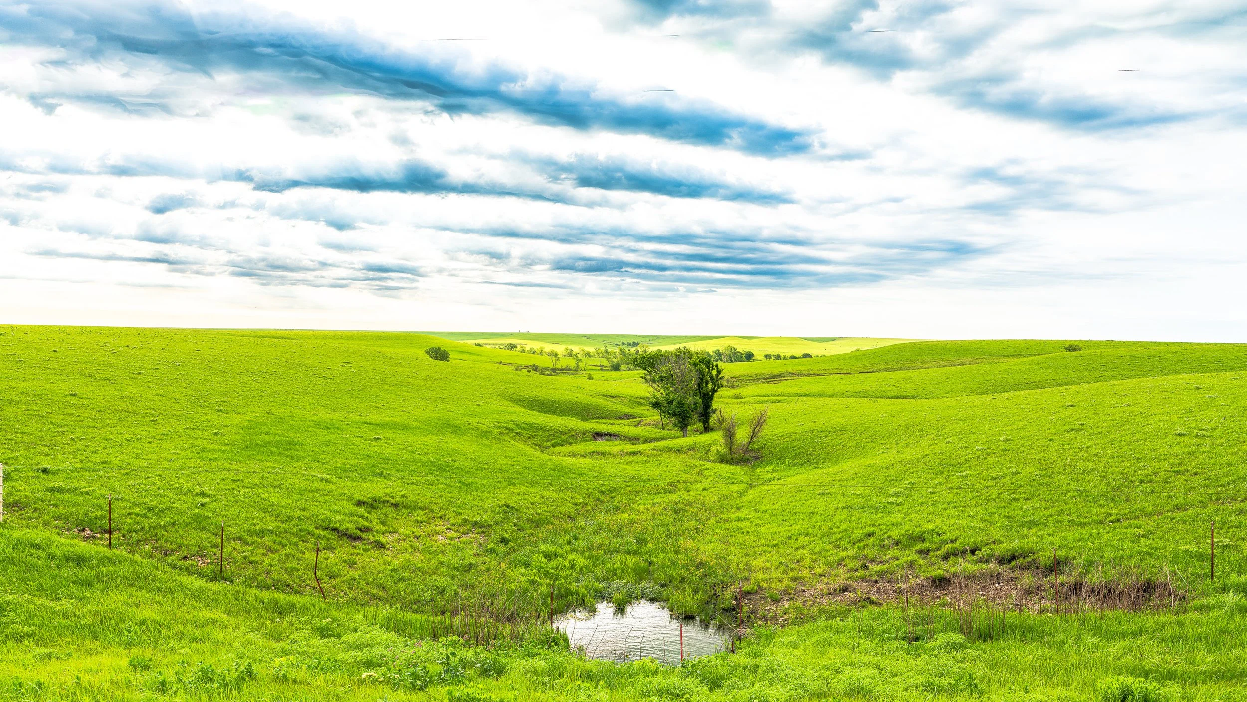 Green rolling hills with a small stream and scattered trees under a partly cloudy sky.